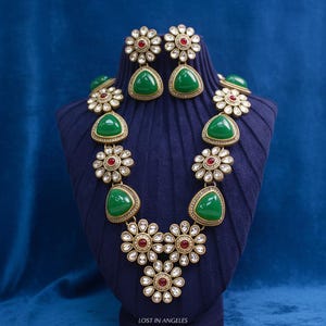 May include: A gold-toned necklace and matching earring set featuring green triangular cabochons and floral-shaped accents with small clear stones and central red gems. The jewelry set is displayed on a dark blue ribbed stand with the text LOST IN ANGELES.
