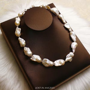May include: A necklace featuring large, irregularly shaped white pearls. The pearls are strung together with a silver clasp. The necklace is displayed on a brown jewelry stand, with the text "LOST IN ANGELES" visible at the bottom.