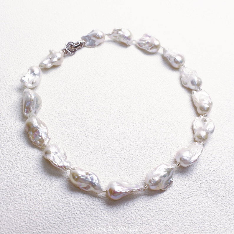 May include: A necklace featuring irregularly shaped, lustrous white pearls strung together with a silver clasp. The pearls have a unique, organic form, reflecting light with a soft sheen. The jewelry is displayed on a white surface.