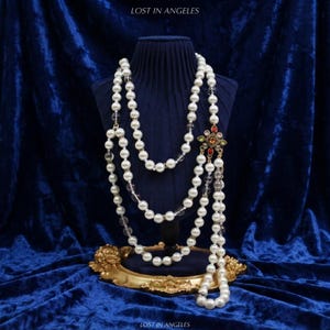 May include: A long multi-strand necklace featuring white faux pearls and clear beads, accented by a central square gold-tone pendant with red, green, and clear crystals. The text LOST IN ANGELES is visible at the top and bottom of the image.