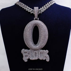Puede incluir: Un collar con colgante plateado con una gran letra "O" y la palabra "BLOCK" en negrita. El colgante cuelga de una gruesa cadena a juego. El texto "LOST IN ANGELES" está en la parte inferior.