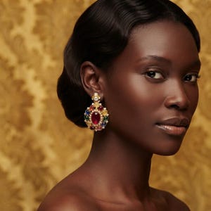 May include: A gold-toned statement earring featuring a large central oval red gemstone surrounded by smaller round blue, green, and yellow cabochon stones, accented with clear crystals in a cluster arrangement on a gold metal base.