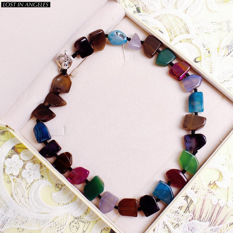 May include: A multi-coloured gemstone necklace featuring various shapes and hues, such as brown, blue, green, and purple. The necklace is presented in a white jewellery box. The text "LOST IN ANGELES" is visible.