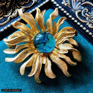 May include: A gold-toned brooch in the shape of a flower with layered petals. The center features a round, blue-green stone with swirling patterns. The brooch is set against a teal background and framed by a black and gold decorative border. The text "LOST IN ANGELS" is at the bottom.