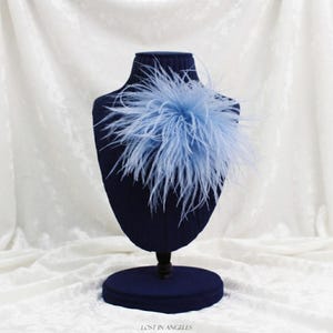May include: A light blue feather accessory attached to a dark navy blue display bust. The feathers are arranged in a circular, voluminous cluster. The text "LOST IN ANGELES" is printed in small, dark letters at the bottom center of the image.