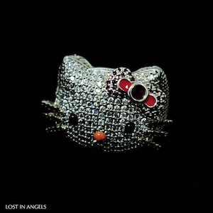 May include: A silver ring in the shape of a cat's head, encrusted with clear gemstones. The cat has black eyes, a small orange nose, and a red bow. The ring is set against a black background. The text "LOST IN ANGELS" is at the bottom.