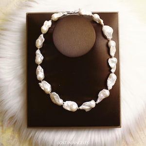 May include: A necklace featuring large, irregularly shaped white pearls strung together. The pearls are displayed on a brown jewelry stand, set against a white fur-like background. The necklace is fastened with a silver clasp.