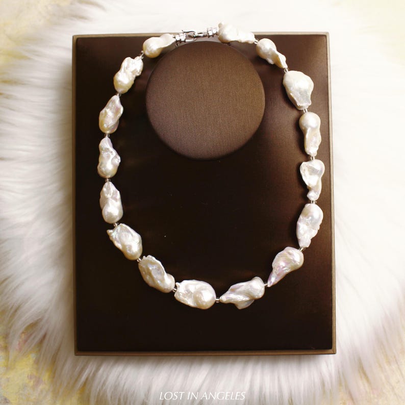 May include: A necklace featuring large, irregularly shaped white pearls strung together. The pearls are displayed on a brown jewelry stand, set against a white fur-like background. The necklace is fastened with a silver clasp.