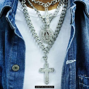 Puede incluir: Primer plano de collares plateados en capas con diseños de cruces y colgantes religiosos. Los collares se llevan sobre una camisa blanca y una chaqueta vaquera. El texto "LOST IN ANGELES" está en la parte inferior.