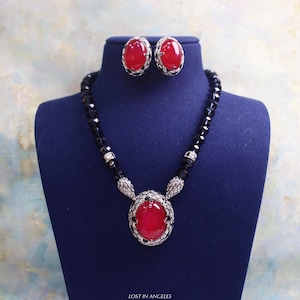 May include: A jewelry set featuring a necklace and matching earrings. The necklace has a large, oval, red gemstone pendant with a black beaded chain. The earrings also have oval red gemstones. The set is displayed on a blue bust.