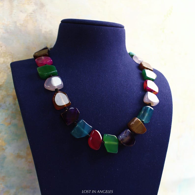 May include: A vibrant gemstone necklace on a blue mannequin. The necklace is made of irregularly shaped stones in shades of green, red, blue, and brown. The text "LOST IN ANGELES" is at the bottom of the image.