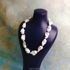 May include: A white pearl necklace displayed on a dark blue mannequin. The necklace features large, irregularly shaped pearls strung together. The background has a colorful, abstract design. The text "LOST IN ANGELES" is visible at the bottom.