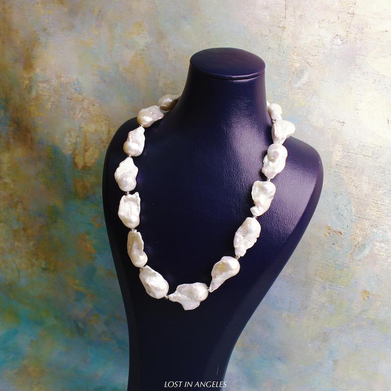 May include: A white pearl necklace displayed on a dark blue mannequin. The necklace features large, irregularly shaped pearls strung together. The background has a colorful, abstract design. The text "LOST IN ANGELES" is visible at the bottom.