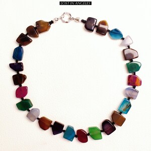 May include: A vibrant necklace composed of multi-coloured, polished gemstones in shades of brown, blue, purple, green, and red. The stones are strung on black cord and fastened with a silver clasp. The text "LOST IN ANGELES" is at the top.