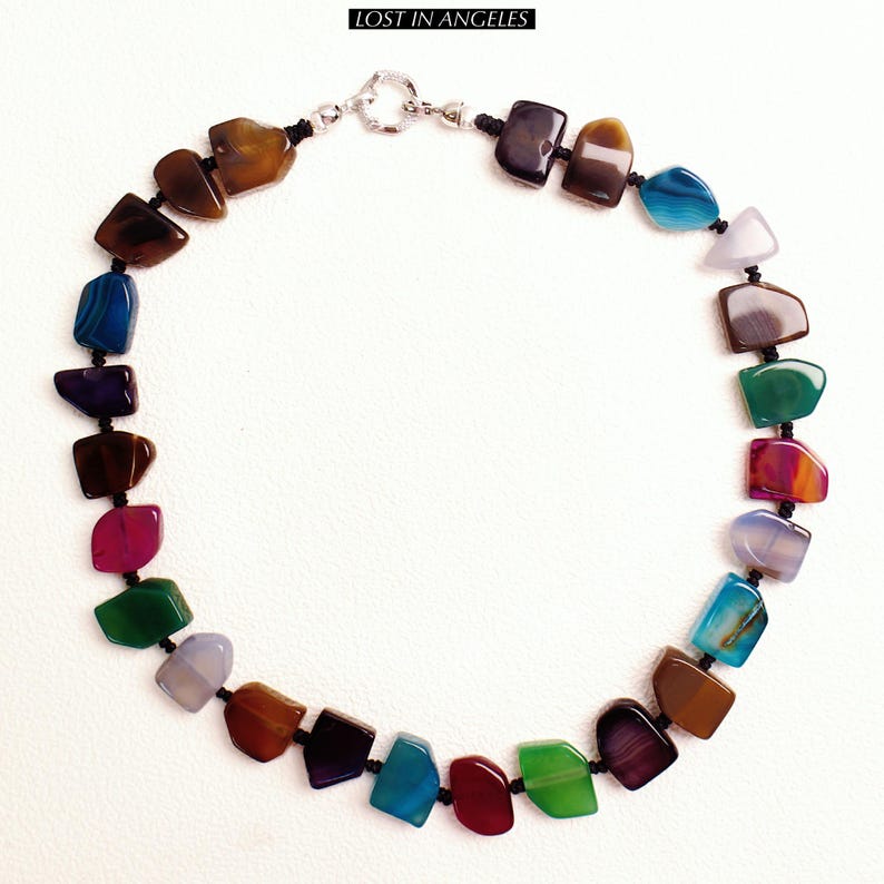 May include: A vibrant necklace composed of multi-coloured, polished gemstones in shades of brown, blue, purple, green, and red. The stones are strung on black cord and fastened with a silver clasp. The text "LOST IN ANGELES" is at the top.