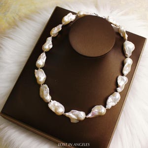 May include: A necklace featuring large, irregularly shaped, off-white pearls strung together with a silver clasp. The pearls have a lustrous sheen and are displayed on a brown surface. The text "LOST IN ANGELES" is visible at the bottom.