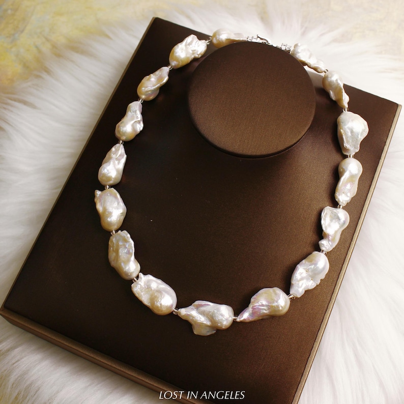 May include: A necklace featuring large, irregularly shaped, off-white pearls strung together with a silver clasp. The pearls have a lustrous sheen and are displayed on a brown surface. The text "LOST IN ANGELES" is visible at the bottom.