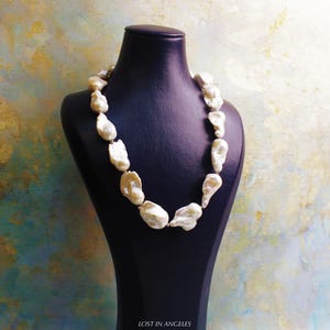 May include: A cream-colored pearl necklace displayed on a black bust. The pearls are irregularly shaped and strung together. The background is a mix of colors, including blue, yellow, and green. The text "LOST IN ANGELES" is at the bottom.