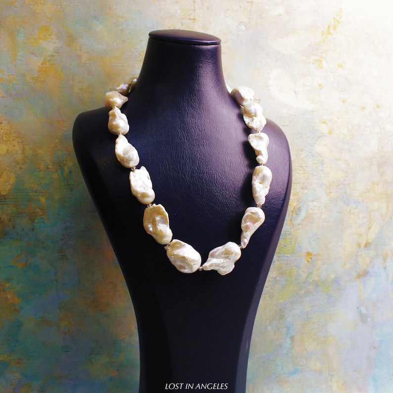 May include: A cream-colored pearl necklace displayed on a black bust. The pearls are irregularly shaped and strung together. The background is a mix of colors, including blue, yellow, and green. The text "LOST IN ANGELES" is at the bottom.