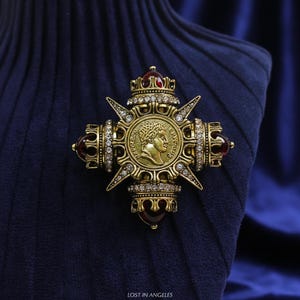 May include: A gold-toned brooch in a cross shape, featuring a central medallion with a profile portrait. The brooch is adorned with clear crystals and red accents, set against a dark blue background. The text "LOST IN ANGELES" is at the bottom.
