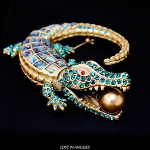May include: Gold-toned alligator brooch with turquoise and abalone accents. The alligator's open mouth holds a gold-colored pearl. The eyes are red. The text "LOST IN ANGELES" is at the bottom.