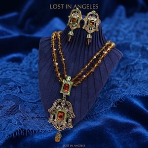 May include: A jewelry set featuring a double-strand necklace and matching earrings with amber-colored crystals, green accents, and metallic settings. The necklace includes a large, ornate pendant with a central amber stone. Text reads LOST IN ANGELES.