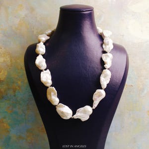 May include: A cream-colored pearl necklace displayed on a dark blue jewelry bust. The necklace features large, irregularly shaped pearls strung together. The background has a textured, multi-colored appearance. The text "LOST IN ANGELES" is at the bottom.