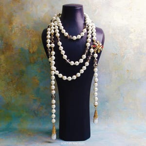 Puede incluir: Un collar de perlas de varias hebras con un cierre decorativo y colgantes colgantes. El collar se exhibe en un maniquí negro. El cierre presenta piedras preciosas rojas, amarillas y verdes. Los colgantes tienen tapas doradas y gotas blancas en forma de lágrima.