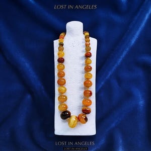 May include: A necklace featuring a single strand of polished, rounded beads in varying shades of yellow, orange, and deep reddish-brown. The beads are strung together, with the largest bead positioned at the center. The text LOST IN ANGELES is visible.