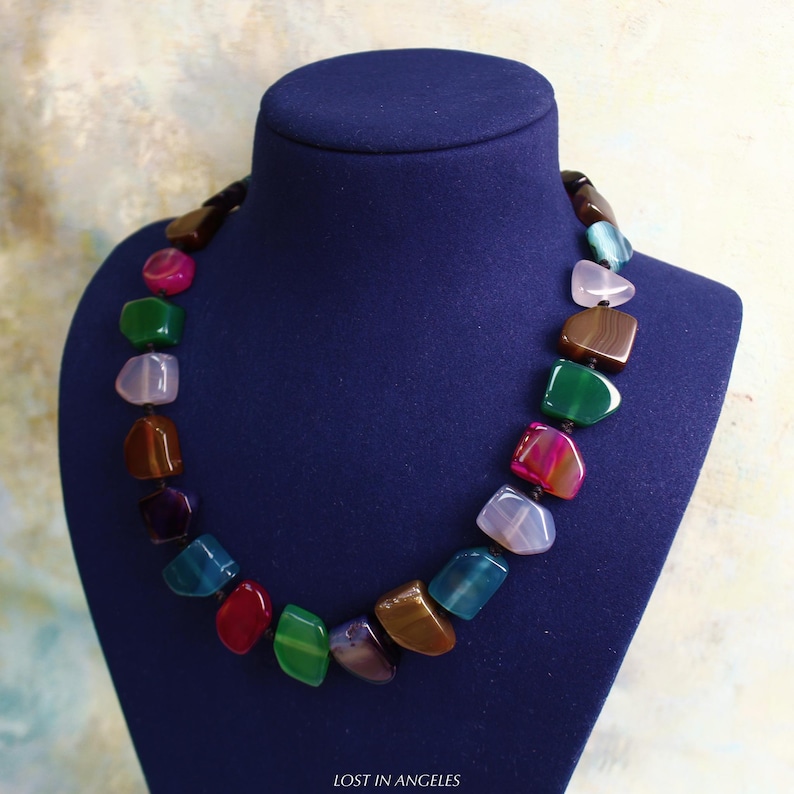 May include: A vibrant gemstone necklace displayed on a blue mannequin. The necklace features a variety of polished stones in shades of red, green, blue, brown, and purple. A striking piece of jewellery.