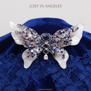 May include: A butterfly-shaped brooch featuring white iridescent wings and a central body encrusted with blue, purple, and clear gemstones. The text LOST IN ANGELES is visible at the top and bottom. The design includes a dangling pear-shaped stone.