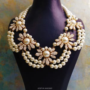 May include: A layered pearl necklace with large, flower-shaped pendants. The necklace features multiple strands of cream-colored pearls, with each flower pendant having a large central pearl surrounded by smaller, white beads and gold-toned accents. The necklace is displayed on a black mannequin.