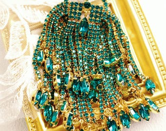 May include: An ornate, emerald green and gold-toned brooch. The brooch features a crown-like top with dangling strands of emerald-colored rhinestones and teardrop-shaped gems. The frame is gold-colored, and the brooch is set against a white lace background.
