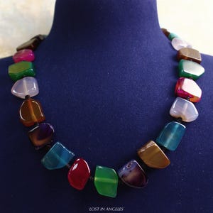 May include: A vibrant gemstone necklace featuring a selection of polished, irregularly shaped stones. The necklace showcases a range of colours, such as red, green, blue, and brown, threaded on a black cord. The text "LOST IN ANGELES" is at the bottom.