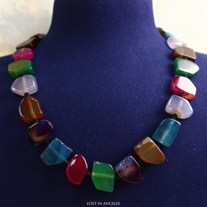 May include: A vibrant gemstone necklace featuring a selection of polished, irregularly shaped stones. The necklace showcases a range of colours, such as red, green, blue, and brown, threaded on a black cord. The text "LOST IN ANGELES" is at the bottom.
