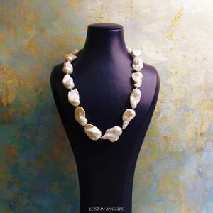 May include: A necklace of large, irregularly shaped, off-white pearls is displayed on a black mannequin. The pearls are strung together, forming a statement piece. The background has a textured, multi-colored appearance with hues of blue, gold, and green.