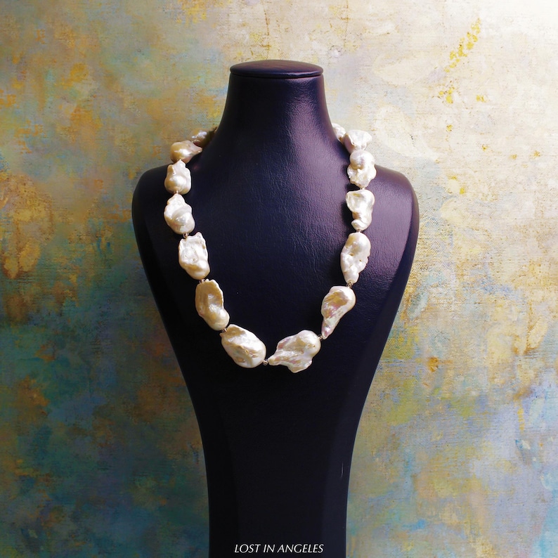 May include: A necklace of large, irregularly shaped, off-white pearls is displayed on a black mannequin. The pearls are strung together, forming a statement piece. The background has a textured, multi-colored appearance with hues of blue, gold, and green.