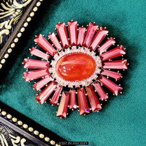 May include: A vibrant, red and pink brooch with a central oval stone surrounded by smaller, sparkling crystals. The brooch features a sunburst design with elongated, rectangular crystals radiating outwards. The text "LOST IN ANGELES" is visible at the bottom.