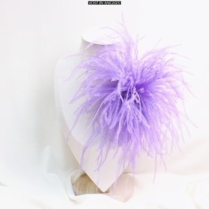 May include: A large, fluffy, lavender-colored feather accent piece, possibly a hair accessory or brooch, is displayed on a white mannequin bust. The piece is centered and has a dense, textured appearance. The text "LOST IN ANGELES" is at the top.