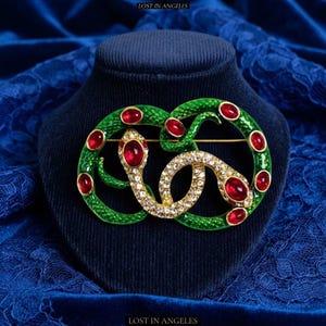 May include: A snake-shaped brooch featuring a green enamelled body with a textured scale pattern, accented by multiple oval-shaped red cabochon stones and a section of clear pavé-set rhinestones. The text LOST IN ANGELES appears at the top and bottom.