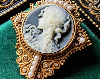 May include: A gold-toned cameo brooch with a white cameo portrait of a woman with a pearl and rhinestone border.