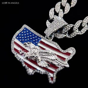 May include: Silver-toned chain necklace with a pendant shaped like the United States map. The pendant features the American flag design with a detailed eagle emblem. The chain and pendant are embellished with sparkling rhinestones. The text "LOST IN ANGELS" is in the top left corner.