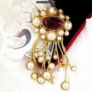 May include: A gold-toned brooch with a large, oval, burgundy-colored stone at its center, surrounded by faux pearls. The brooch has multiple strands of gold chains with pearl drops, and the text "LOST IN ANGELS" is visible.