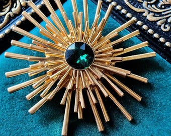 May include: A gold-toned brooch in the shape of a sun with a large green gemstone in the center.