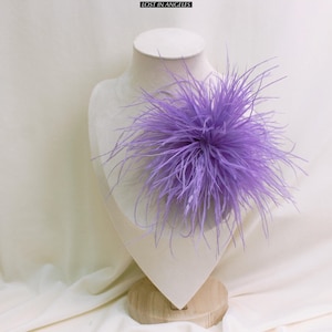 May include: A lavender feather accent piece, displayed on a mannequin bust. The accessory is a cluster of long, wispy feathers, creating a soft, textured appearance. The text "LOST IN ANGELES" is at the top of the image. The bust is set against a neutral background.
