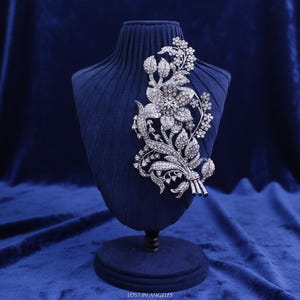 Puede incluir: Un broche floral plateado con brillantes cristales transparentes, exhibido sobre un busto de terciopelo azul. El broche presenta una flor grande, hojas y pequeños detalles florales, creando un diseño elegante y detallado. El texto "LOST IN ANGELES" es visible.