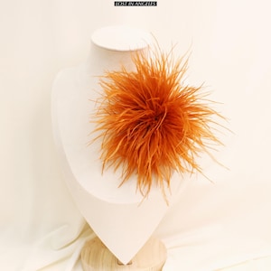 May include: An orange feather accent piece, possibly a brooch or hair accessory, displayed on a white mannequin bust. The accessory is a dense cluster of long, spiky feathers. The brand name "LOST IN ANGELS" is visible at the top.