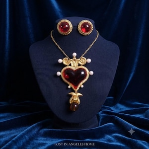 May include: A gold-tone necklace and earring set featuring a large heart-shaped pendant with a deep red center, accented by small white pearls and a dangling gold charm. The matching stud earrings feature round, deep red stones in gold settings.
