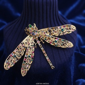 May include: A gold-toned dragonfly brooch featuring an intricate openwork metal design encrusted with numerous small, multi-colored rhinestones in shades of red, blue, green, and yellow. The text LOST IN ANGELES is printed at the bottom center.