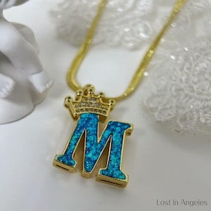 May include: Gold chain necklace with a blue and gold letter M pendant. The letter M is encrusted with blue glitter and topped with a gold crown with clear rhinestones.
